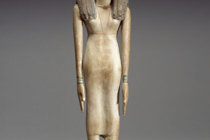While she wears a fashionable wig, this woman's own close-cropped hair can be seen at the forehead as well as on the sides of her neck. Her graceful proportions are set off by her elongated arms and emphasized hands and feet that are characteristic of Middle Kingdom sculptures. As is often the case, this statue has carefully indicated fingernails and toenails. There is a diadem running all over this statuette's wig from the middle. The wig seems to be attached to the body as well as the arms. The woman has armlets and anklets painted in green color with black lines. It seems that the wooden body was painted. Some white colors remain on the fingers. She is dressed in the traditional linen sheath with shoulder straps. Hair in pigtails ending in short curls bound with band at ear level, hanging beside neck and down back. Her wig was made by securing human hair to a mesh foundation by means of beeswax and resin. The large wig covers the ears, the brownish color maybe was due to the fading black color that was before. The legs are side by side. Her arms are at her sides. Her hands are extended flat. Her feet are glued to the block base. Her eyelid is not fully extended to the end of face, as well as the thick eyebrows. The black canthus is wide. She has a round face and a long neck. She is wearing a necklace with large central bead. On the back, the piece is partially broken from above the left leg. There is a brownish line on the back, extending from front, under the waist.