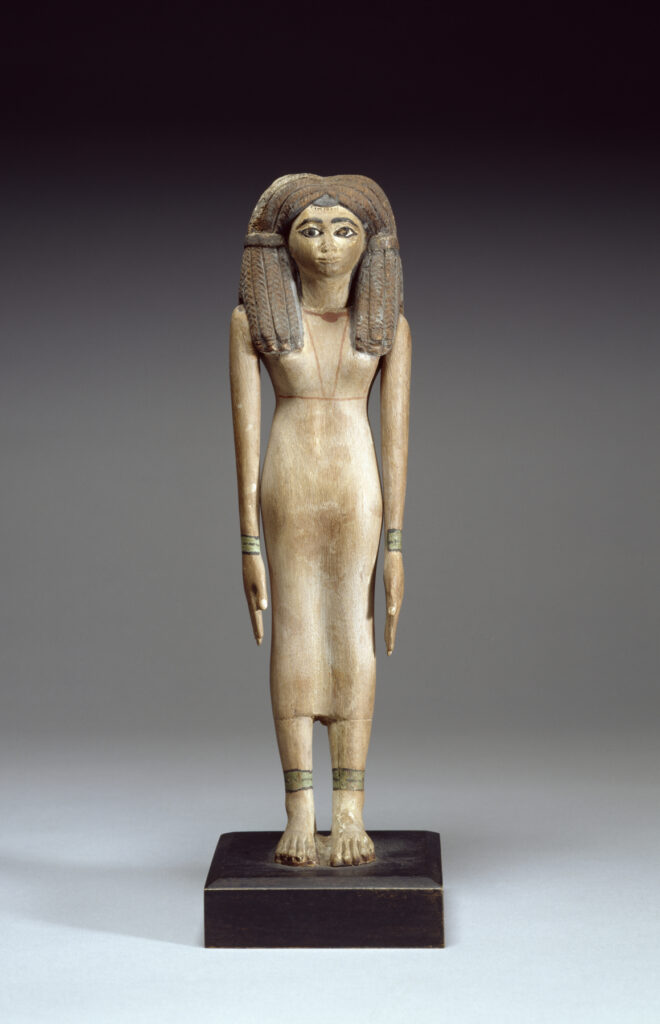 While she wears a fashionable wig, this woman's own close-cropped hair can be seen at the forehead as well as on the sides of her neck. Her graceful proportions are set off by her elongated arms and emphasized hands and feet that are characteristic of Middle Kingdom sculptures. As is often the case, this statue has carefully indicated fingernails and toenails. There is a diadem running all over this statuette's wig from the middle. The wig seems to be attached to the body as well as the arms. The woman has armlets and anklets painted in green color with black lines. It seems that the wooden body was painted. Some white colors remain on the fingers. She is dressed in the traditional linen sheath with shoulder straps. Hair in pigtails ending in short curls bound with band at ear level, hanging beside neck and down back. Her wig was made by securing human hair to a mesh foundation by means of beeswax and resin. The large wig covers the ears, the brownish color maybe was due to the fading black color that was before. The legs are side by side. Her arms are at her sides. Her hands are extended flat. Her feet are glued to the block base. Her eyelid is not fully extended to the end of face, as well as the thick eyebrows. The black canthus is wide. She has a round face and a long neck. She is wearing a necklace with large central bead. On the back, the piece is partially broken from above the left leg. There is a brownish line on the back, extending from front, under the waist.